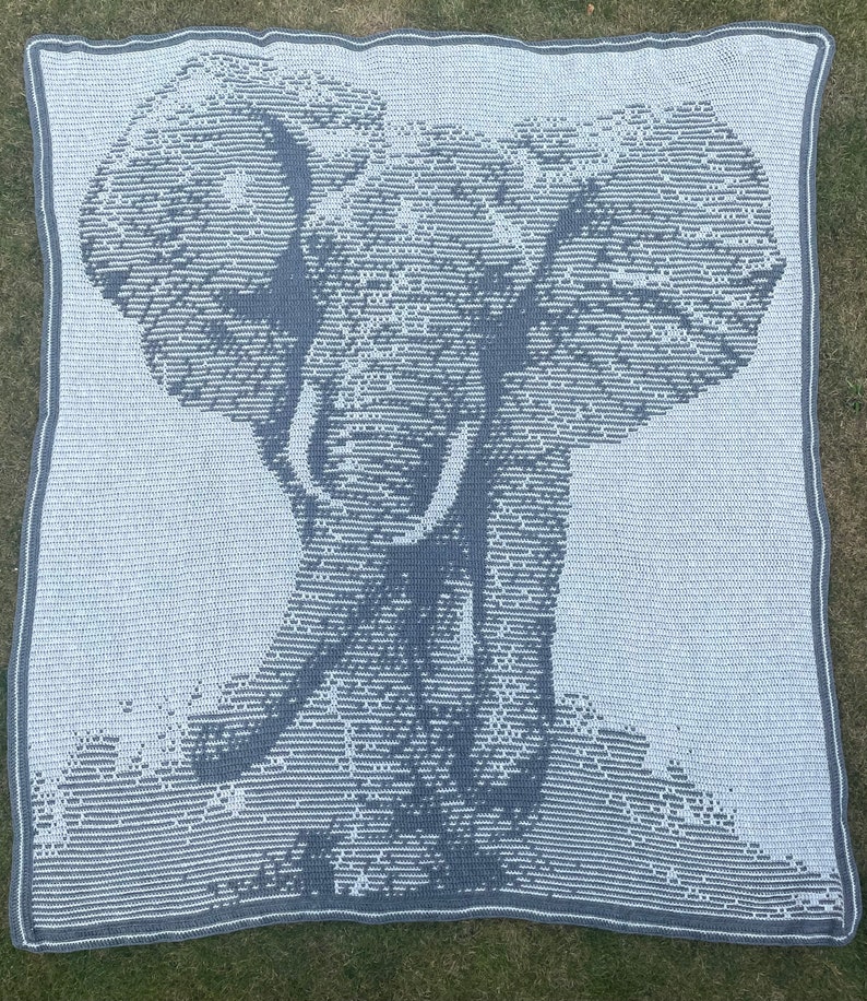 Puede incluir: Una manta de ganchillo con la imagen de un elefante en tonos grises y blancos. El dise&ntilde;o del elefante es detallado, con la trompa y los colmillos bien visibles. La manta tiene un borde gris y est&aacute; colocada sobre hierba. Dimensiones: aprox. 150 cm x 150 cm.