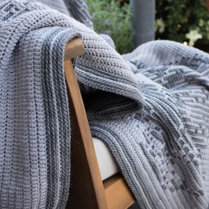Puede incluir: Una manta de ganchillo gris y blanca sobre una silla de madera. La manta presenta varios patrones texturizados y un borde a rayas. La paleta de colores es neutra, con tonos de gris y blanco, creando una est&eacute;tica acogedora e invitadora.