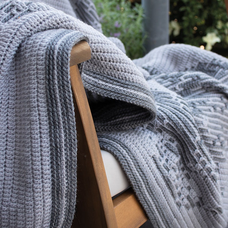 Puede incluir: Una manta de ganchillo gris y blanca sobre una silla de madera. La manta presenta varios patrones texturizados y un borde a rayas. La paleta de colores es neutra, con tonos de gris y blanco, creando una est&eacute;tica acogedora e invitadora.
