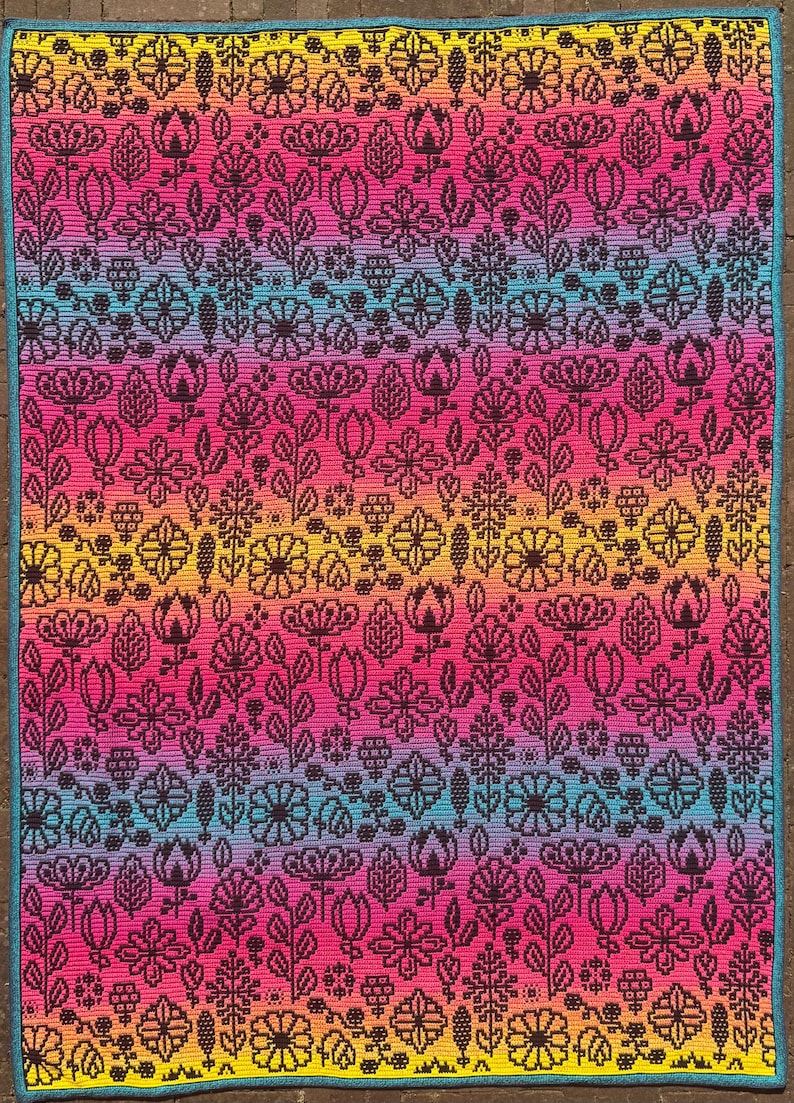 May include: A colorful crochet blanket with a rainbow gradient pattern featuring black floral designs. The blanket is made with a variety of stitches and textures.