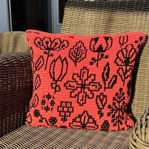 May include: A coral-colored pillow with a black geometric floral pattern. The pillow is sitting on a wicker chair.