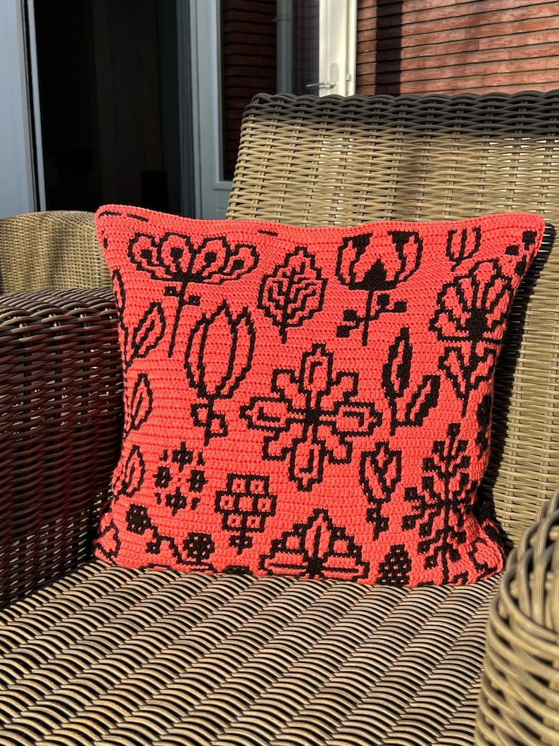 May include: A coral-colored pillow with a black geometric floral pattern. The pillow is sitting on a wicker chair.