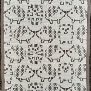 Op de afbeelding: Een wit en grijs gehaakte deken met een herhalend patroon van gepixelde egels. De deken heeft een grijze rand. Deze handgemaakte deken is ideaal voor een baby- of kinderkamer.