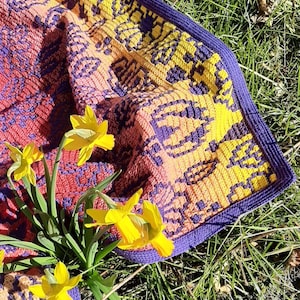 May include: A crocheted blanket with a purple, yellow, and orange geometric pattern. The blanket is lying on a bed of green grass with yellow daffodils.