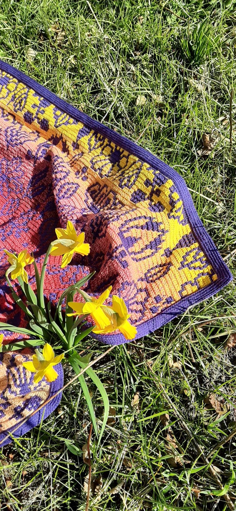 May include: A crocheted blanket with a purple, yellow, and orange geometric pattern. The blanket is lying on a bed of green grass with yellow daffodils.