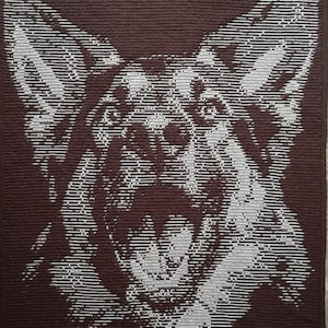 Op de afbeelding: Een bruin en wit gehaakt wandkleed met daarop een Duitse herder met zijn bek open. De vacht van de hond is gemaakt van wit en bruin garen, de achtergrond is effen bruin.