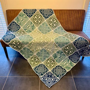 Op de afbeelding: Een gehaakt deken in groen, wit en blauw met een geometrisch patroon. Het deken ligt over een bruine bank.