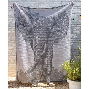 Puede incluir: Una manta rectangular gris y blanca con un dise&ntilde;o detallado de la cabeza de un elefante. La manta se sostiene contra una pared de ladrillos blancos, mostrando el intrincado dise&ntilde;o y la textura.