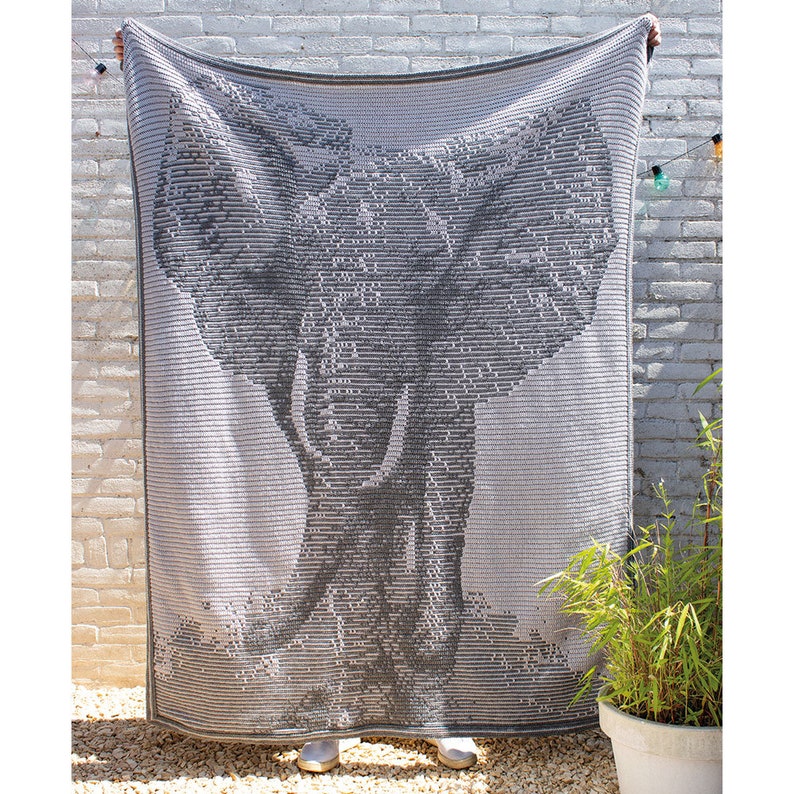 Puede incluir: Una manta rectangular gris y blanca con un dise&ntilde;o detallado de la cabeza de un elefante. La manta se sostiene contra una pared de ladrillos blancos, mostrando el intrincado dise&ntilde;o y la textura.