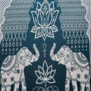 May include: A teal blue tapestry featuring a white design of two elephants facing each other with a lotus flower between them. The elephants have intricate patterns on their bodies and the lotus flower has a detailed design. The background is a repeating pattern of diamonds.