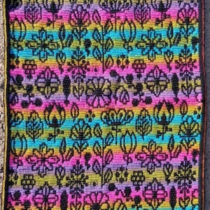 May include: A colorful crocheted square with a floral pattern in shades of pink, purple, blue, yellow, and green. The design features various flowers and leaves in a repeating pattern.