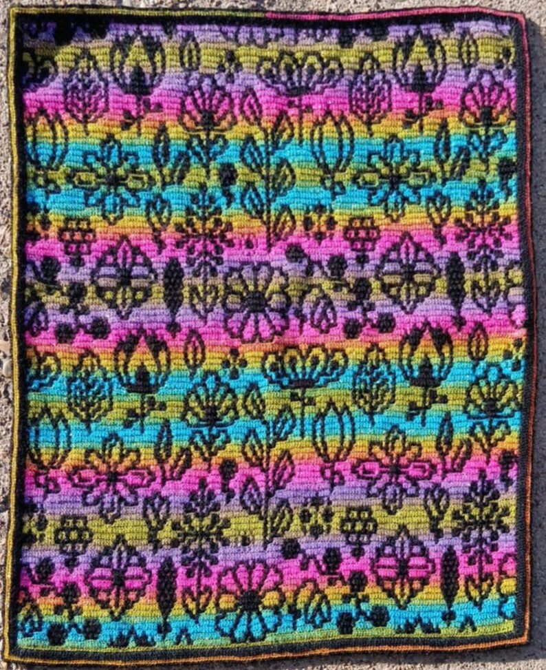 May include: A colorful crocheted square with a floral pattern in shades of pink, purple, blue, yellow, and green. The design features various flowers and leaves in a repeating pattern.
