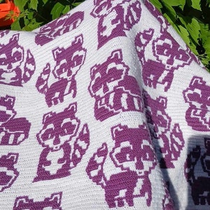 May include: A white crocheted blanket with a purple pattern of raccoons. The blanket is folded and draped over a chair.