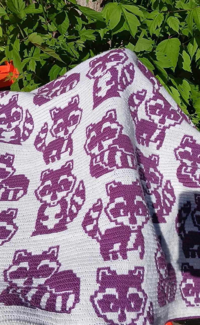 May include: A white crocheted blanket with a purple pattern of raccoons. The blanket is folded and draped over a chair.