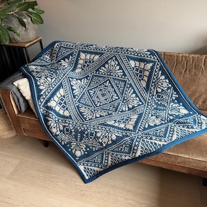 Op de afbeelding: Een blauw-wit patroon deken is gedrapeerd over een bruine leren bank. De deken heeft een geometrisch ontwerp met bloem- en stervormige motieven. De rand van de deken is effen blauw.