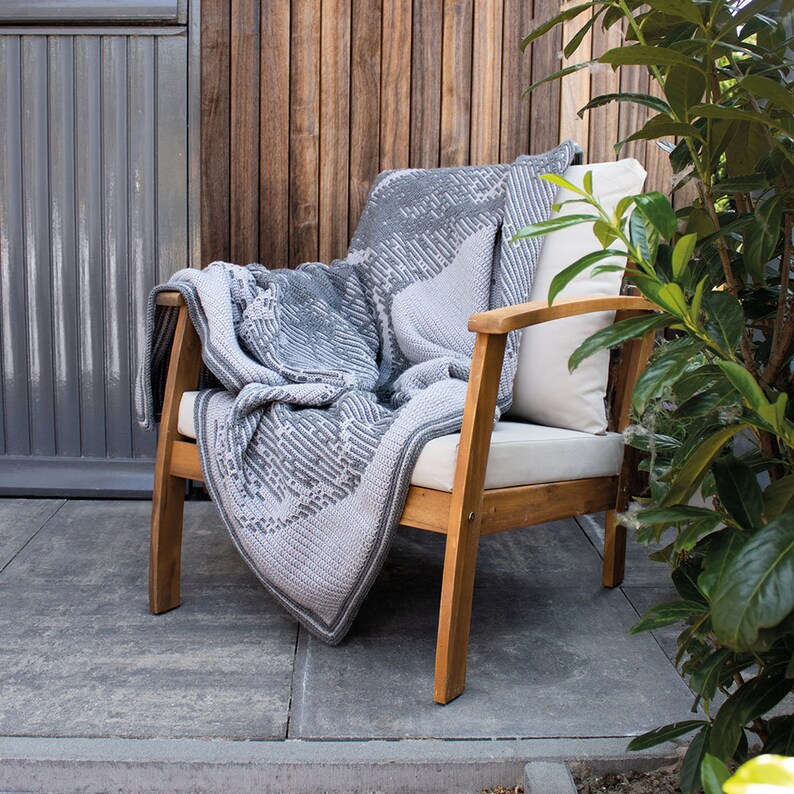 Puede incluir: Un sill&oacute;n de madera con un coj&iacute;n claro y una manta gris tejida con un dise&ntilde;o geom&eacute;trico. La manta tiene un borde gris oscuro. Una almohada a juego descansa sobre la silla. La escena est&aacute; ambientada al aire libre con una pared de madera y vegetaci&oacute;n.