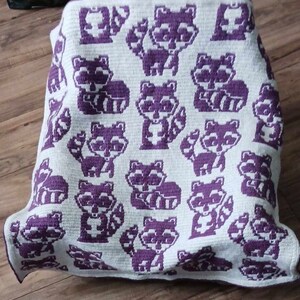 May include: A crocheted blanket with a white background and purple raccoon designs. The blanket is folded and lying on a wooden floor.