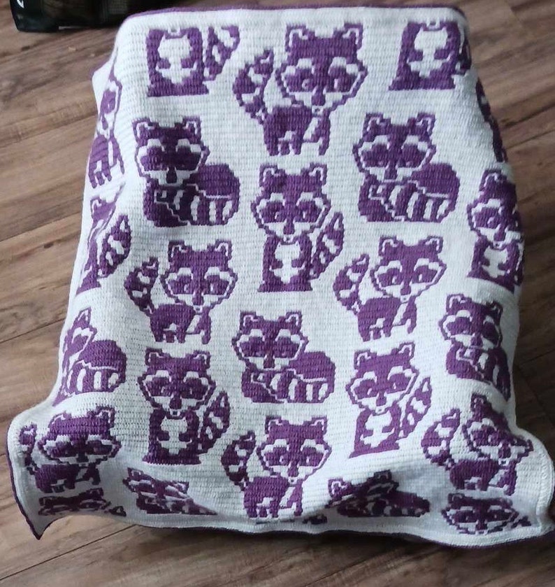 May include: A crocheted blanket with a white background and purple raccoon designs. The blanket is folded and lying on a wooden floor.