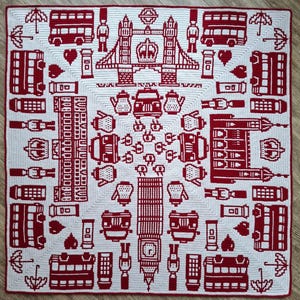 Op de afbeelding: Een vierkante gehaakte deken met een witte achtergrond en rode patronen van Londense bezienswaardigheden, waaronder Tower Bridge, Big Ben, dubbeldekkerbussen, telefooncellen, wachters, theepotten, paraplu's en kronen in een symmetrisch ontwerp.