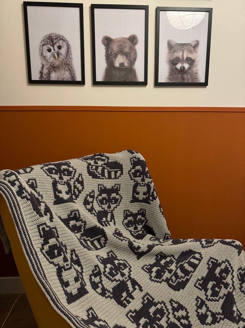 May include: A gray and black crocheted blanket with a pixelated design of raccoons. The blanket is draped over a yellow chair in front of a wall with three framed animal prints.