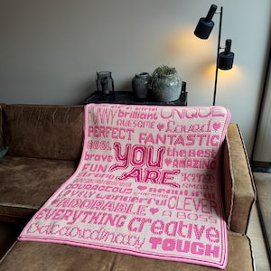 Pode incluir: Um cobertor de crochê rosa e creme com as palavras "YOU ARE" em letras rosa grandes. O cobertor é adornado com palavras positivas como "FUNNY", "BRAVE" e "CLEVER". Está sobre um sofá castanho.