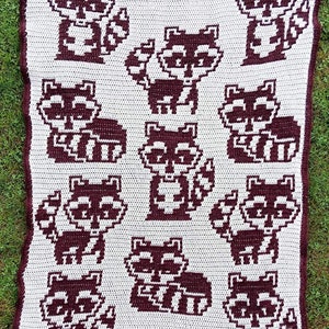May include: A white crocheted blanket with a brown pixelated pattern of raccoons. The blanket features multiple raccoons in different poses.