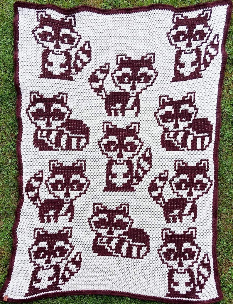 May include: A white crocheted blanket with a brown pixelated pattern of raccoons. The blanket features multiple raccoons in different poses.