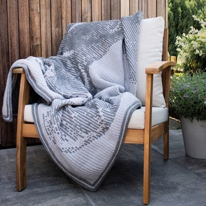 Puede incluir: Una manta y un coj&iacute;n de ganchillo grises y blancos sobre una silla de madera. La manta presenta un patr&oacute;n geom&eacute;trico y un borde en contraste. La silla est&aacute; en un patio con una valla de madera al fondo.