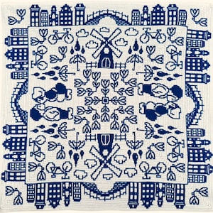 Può includere: Un quadrato di uncinetto bianco con un design blu che presenta mulini a vento, biciclette, tulipani e case. Il design è simmetrico e ha un tema olandese.