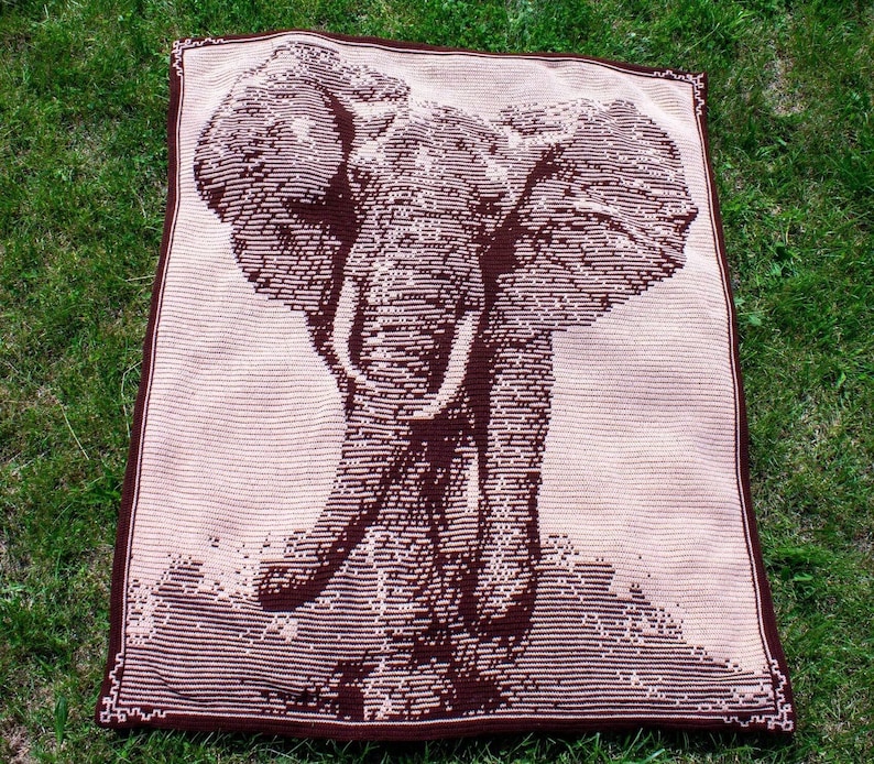 Puede incluir: Una manta tejida marr&oacute;n y beige con un dise&ntilde;o detallado de un elefante. El elefante se representa en un estilo lineal, con su trompa, colmillos y orejas claramente visibles. La manta tiene un borde marr&oacute;n oscuro y est&aacute; sobre hierba verde. Medidas en cent&iacute;metros.