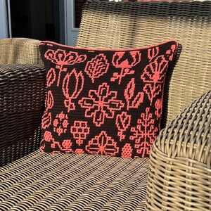 May include: A black and coral crocheted pillow with a floral pattern sits on a wicker chair.