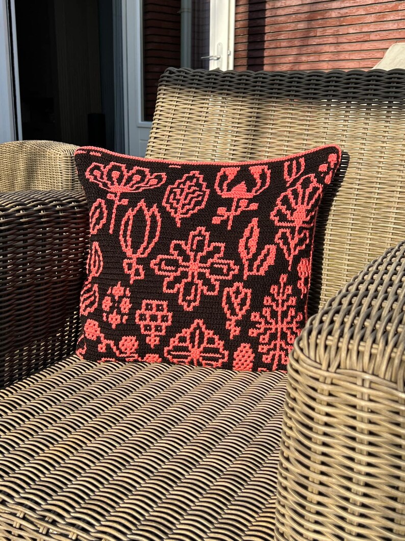 May include: A black and coral crocheted pillow with a floral pattern sits on a wicker chair.