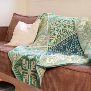 Puede incluir: Una manta de crochet verde y azul con un patrón de copo de nieve está colocada sobre un sofá de pana marrón.