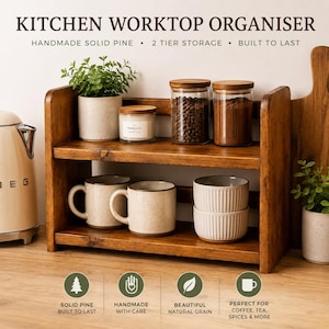 May include: A two-tier wooden kitchen worktop organiser made of solid pine. The top shelf holds a small potted plant, a vanilla candle, and two glass jars filled with coffee beans and grounds. The bottom shelf holds two speckled mugs and two ribbed bowls.