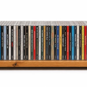 May include: A light brown wooden storage rack holding a row of numerous music CDs in their plastic jewel cases. The rack features a simple, open-ended design with side panels and a base, displaying a variety of colourful album spines side by side.