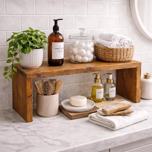 May include: A rustic wooden countertop shelf holding a green plant in a white pot, an amber glass hand soap dispenser, a clear glass jar filled with white cotton balls, and a woven basket containing white rolled towels and washcloths.