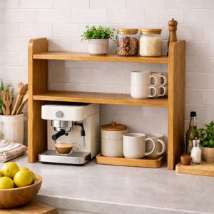 May include: A two-tier wooden countertop shelf holding a white espresso machine, a glass cup with coffee, two stacked white mugs, a lidded ceramic canister, and two glass jars filled with food items, with a small potted green plant on top.