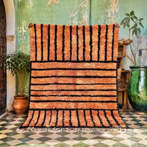 Peut inclure: Un tapis rectangulaire à rayures orange et noires avec une surface texturée. Le tapis présente une série de rayures noires horizontales et des rayures noires verticales en haut et en bas. Le tapis est présenté sur un fond vert et blanc.