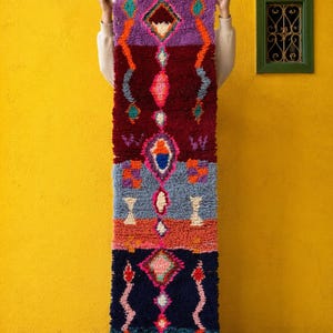 Puede incluir: Una alfombra rectangular vertical con un diseño geométrico vibrante. La alfombra presenta una mezcla de colores que incluyen morado, burdeos, naranja, gris y azul marino. El diseño incluye formas de diamante y ovaladas, con un borde con flecos en la parte inferior.