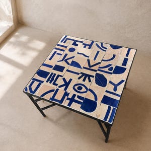 Peut inclure: Une table basse carrée avec un cadre en métal noir et un plateau composé de carreaux de céramique beige clair. Les carreaux présentent des formes géométriques, des lignes et des symboles abstraits bleu foncé peints à la main en grille.