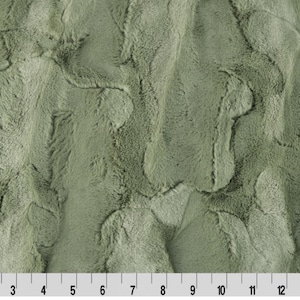 May include: A close-up of a soft, plush fabric in a muted green color. The fabric has a subtle marbled pattern and a fuzzy texture.