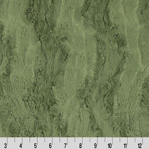 May include: Close-up of a sage green faux fur fabric with a textured, wavy pattern. The fabric has a soft, plush appearance. A ruler is at the bottom, showing measurements in inches.