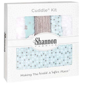 May include: A white box labeled "Cuddle Kit" with a window revealing fabric swatches. The fabrics include white, light blue with gray polka dots, and a textured tan material. The "Shannon Fabrics" logo is visible, along with the tagline "Making The World A Softer Place."