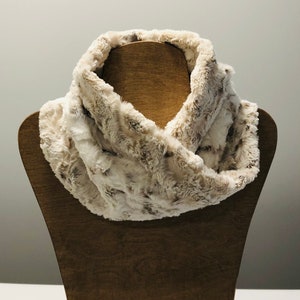 May include: A soft, faux fur infinity scarf in a light brown and white color. The scarf is made of a plush, fuzzy material and is perfect for keeping warm in the winter.