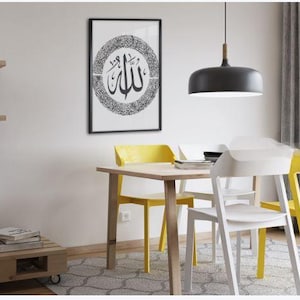 K&ouml;nnte beinhalten: Ein gerahmtes Schwarz-Wei&szlig;-Kunstwerk mit arabischer Kalligraphie h&auml;ngt &uuml;ber einem Holztisch mit gelben und wei&szlig;en St&uuml;hlen. Eine schwarze Pendelleuchte h&auml;ngt &uuml;ber dem Tisch. Der Raum hat ein modernes, minimalistisches Design.
