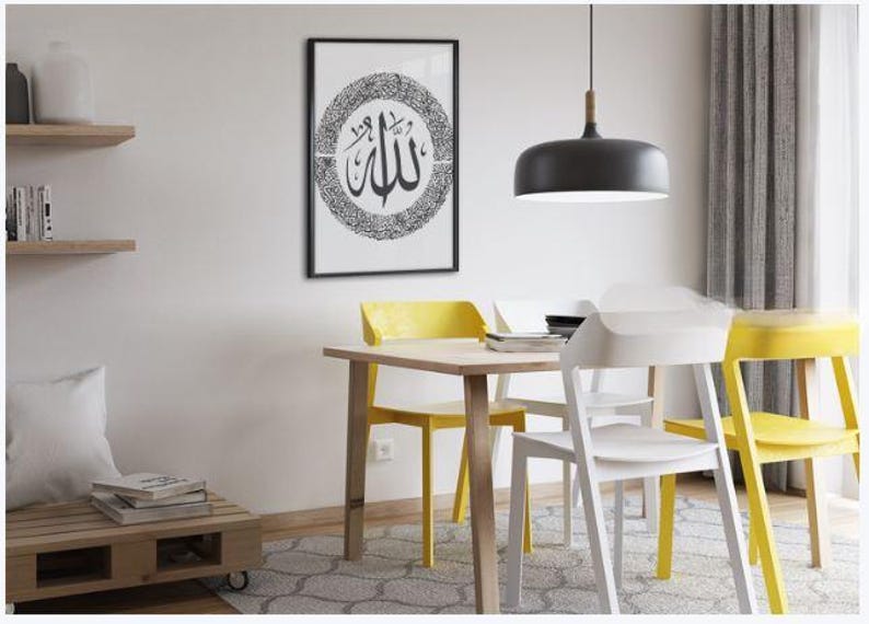 K&ouml;nnte beinhalten: Ein gerahmtes Schwarz-Wei&szlig;-Kunstwerk mit arabischer Kalligraphie h&auml;ngt &uuml;ber einem Holztisch mit gelben und wei&szlig;en St&uuml;hlen. Eine schwarze Pendelleuchte h&auml;ngt &uuml;ber dem Tisch. Der Raum hat ein modernes, minimalistisches Design.