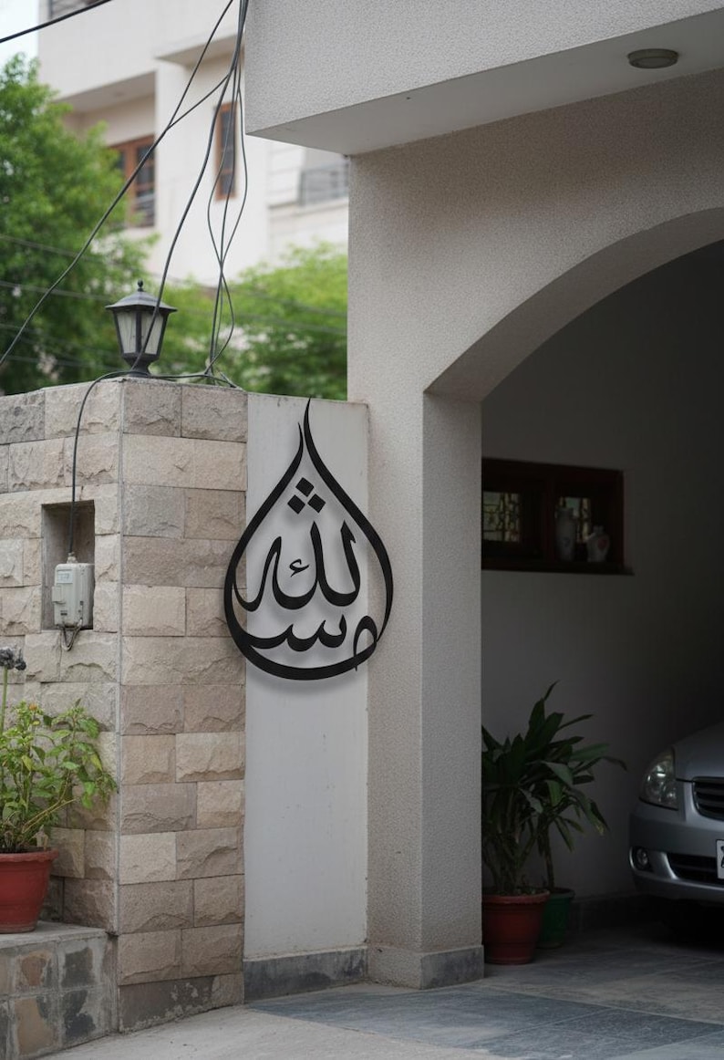 K&ouml;nnte beinhalten: Ein schwarzes Metall-Wandkunstwerk in Tropfenform mit arabischer Kalligraphie. Das Kunstwerk ist an einem wei&szlig;en Pfeiler neben einer Steinmauer angebracht. Eine traditionelle Au&szlig;enlampe ist an der Steinmauer montiert.