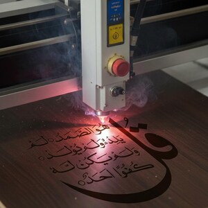 K&ouml;nnte beinhalten: Eine Lasergravurmaschine graviert arabische Kalligraphie auf eine Holzoberfl&auml;che. Der Laser emittiert einen roten Strahl, der Rauch erzeugt, w&auml;hrend er die komplizierte Schrift graviert. Das Holz hat einen warmen, braunen Farbton, und der Text ist dunkel.