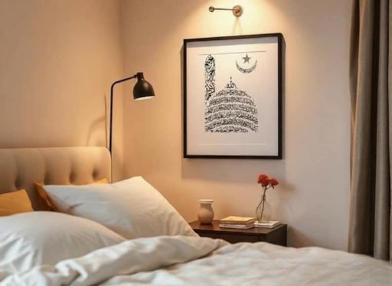 K&ouml;nnte beinhalten: Ein Schlafzimmer mit einem Bett mit wei&szlig;er Bettw&auml;sche und Kissen. Ein gerahmter schwarz-wei&szlig;er islamischer Kunstdruck h&auml;ngt &uuml;ber dem Bett. Ein kleiner Nachttisch mit einer Vase mit Blumen und einer Lampe.