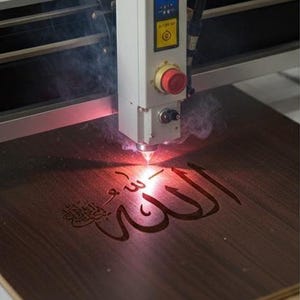 K&ouml;nnte beinhalten: Eine Lasergravurmaschine graviert arabische Kalligraphie auf eine dunkle Holzoberfl&auml;che. Die wei&szlig;en und grauen Komponenten der Maschine sind sichtbar, mit einem roten Not-Aus-Knopf. Der Gravurprozess erzeugt Rauch und ein hellrotes Licht.